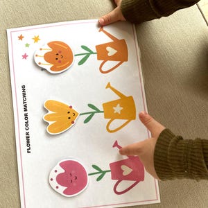 K&ouml;nnte beinhalten: Ein Blumenfarben-Zuordnungsspiel mit drei Blumen und drei Gie&szlig;kannen. Die Blumen sind orange, gelb und rosa. Die Gie&szlig;kannen haben die gleichen Farben wie die Blumen. Das Spiel ist mit "Blumenfarben-Zuordnung" beschriftet.