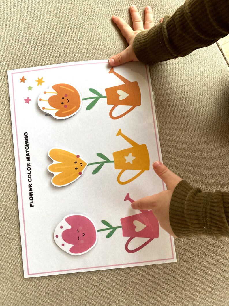 K&ouml;nnte beinhalten: Ein Blumenfarben-Zuordnungsspiel mit drei Blumen und drei Gie&szlig;kannen. Die Blumen sind orange, gelb und rosa. Die Gie&szlig;kannen haben die gleichen Farben wie die Blumen. Das Spiel ist mit "Blumenfarben-Zuordnung" beschriftet.