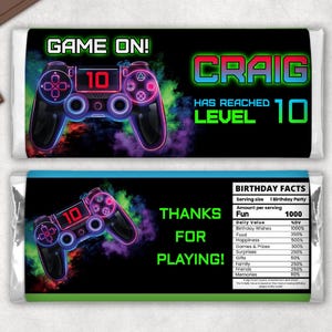 Pode incluir: Um design de embalagem de barra de chocolate imprimível com um gráfico de controlador de videogame verde neon, azul e vermelho. O texto "GAME ON!" está em verde, e o texto "CRAIG HAS REACHED LEVEL 10" está em verde, azul e vermelho. A embalagem também inclui uma seção com o texto "THANKS FOR PLAYING!" e uma tabela com fatos de aniversário.