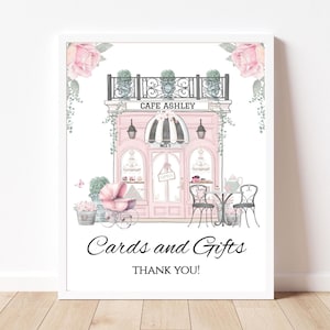 Op de afbeelding: Waterverfillustratie van een roze café met de tekst "Cafe Ashley" en "Kaarten en cadeaus". De afbeelding toont een kinderwagen, bloemaccenten en een decoratieve luifel. De woorden "Dank u!" zijn ook opgenomen.