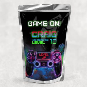 May include: A silver foil pouch with a black background and a neon green and pink video game controller graphic. The text "GAME ON!" is in green, "CRAIG" is in pink, "HAS REACHED LEVEL 10" is in green, and "LEVEL UP" is in white.