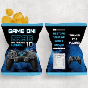 以下が含まれることがあります： 「GAME ON! CRAIG HAS REACHED LEVEL 10」の文字が入った黒と青のチップス袋。ゲームコントローラーのグラフィックと「LEVEL UP」の文字が特徴です。袋の裏面には「RESTORE YOUR HP WITH A SNACK!」と「THANKS FOR PLAYING!」と書かれています。