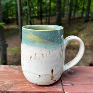 May include: A ceramic mug featuring a two-tone glaze with a smooth teal and yellow upper section and a textured off-white lower body. The base and handle display subtle brown speckles and vertical drip-like markings across the surface.