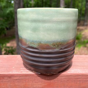 May include: A handmade ceramic mug with a cylindrical shape. The top half is a light green color, transitioning to a brown band with a textured, speckled appearance. The bottom half is a dark brown with horizontal ridges.