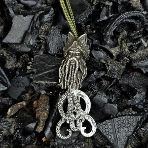May include: A silver octopus pendant with a bronze pirate head and beard hanging from a green cord. The pendant is on a black background.