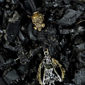 May include: A silver and gold pendant featuring a winged figure with a detailed design. The pendant is hanging from a black cord with a gold gas mask charm.