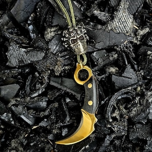 Puede incluir: Un collar con colgante que presenta un cuchillo karambit dorado con una hoja curva y un mango de madera oscura. El collar también incluye un dije en forma de calavera y un cordón verde. El fondo es una superficie oscura y texturizada.