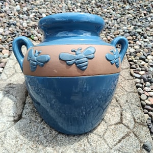 May include: A blue ceramic planter with a wide base and a narrower top. The planter features two handles and a band of tan-coloured ceramic with three embossed bee designs. The planter is sitting on a stone surface.