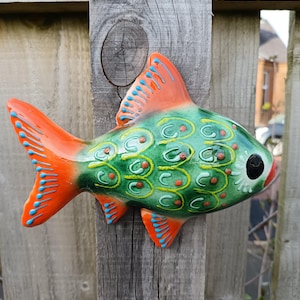 May include: A colourful ceramic fish sculpture, mounted on a weathered wooden fence. The fish has a green body with orange fins and blue detailing. The fish features a detailed scale pattern and a black eye.