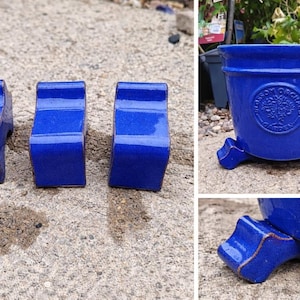 May include: A collection of three cobalt blue ceramic plant pot feet and a matching blue ceramic plant pot. The pot features a round emblem with the words "Blossom Orchard Garden". The pot has three feet.
