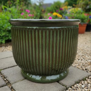 May include: A large, green ceramic planter with vertical ribbed detailing. The planter has a wide, slightly flared rim and sits on a rounded base. The glossy finish gives it a polished look, suitable for outdoor use. The background shows a garden setting.