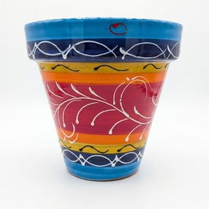 May include: A brightly coloured ceramic plant pot. It has horizontal bands of blue, orange, red, and yellow, with white floral patterns and decorative lines. The top rim is solid blue. The pot is approximately 15 cm tall.