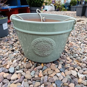 May include: A sage green ceramic hanging planter with a rope handle. The planter has a round emblem that reads "Blossom Orchard Garden". The planter is sitting on a bed of small, colorful stones. The planter is designed for outdoor use.