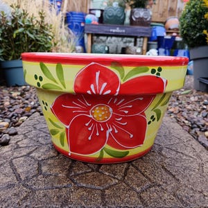 May include: A hand-painted ceramic flower pot featuring a large red flower on a yellow base. The rim is red, with green leaves and dots. This decorative pot is suitable for indoor or outdoor use. The pot's diameter is approximately 25 cm.