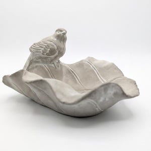 May include: A grey concrete bird bath shaped like a large leaf. A small bird statue sits on the edge of the leaf. The bird bath is a light grey colour and has a textured surface.