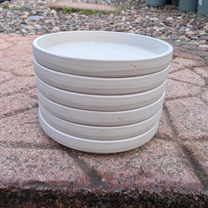 May include: A stack of six light grey ceramic plant saucers. The saucers are round with a slightly raised edge and sit on a red brick surface. The background shows other plant pots and gardening supplies.