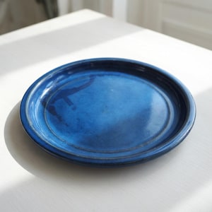 May include: A round, cobalt blue ceramic plate with a slightly raised rim. The plate has a glossy finish and a subtle, textured surface. The plate is sitting on a white surface.