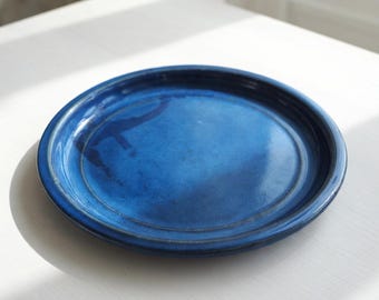 May include: A circular ceramic plate featuring a deep, glossy cobalt blue glaze with subtle variations in colour intensity across the surface. The plate has a slightly raised rim and a smooth, reflective finish that catches the light.