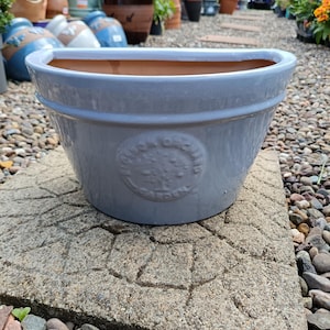 May include: A large, pale grey ceramic plant pot with a wide, oval opening. The pot has a textured surface and a decorative emblem on the front. The interior is terracotta. Suitable for outdoor gardening.