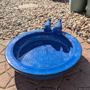 May include: A circular, blue ceramic bird bath with two blue bird ornaments on the edge. The bird bath is filled with water and is placed on a brick patio. This is a decorative garden accessory, perfect for attracting birds.