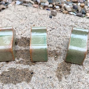 May include: Three ceramic cabinet pulls with a rectangular shape. The pulls have a green and brown colour scheme with a glossy finish. The pulls are arranged on a concrete surface.
