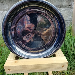 May include: A glazed ceramic bowl in dark blue, with a wide, shallow design. The bowl, supported by a wooden stand, has a glossy finish and a swirling interior pattern of purple, brown, and gold. The bowl's diameter is approximately 30 cm.