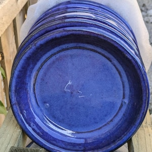 May include: A pile of deep blue ceramic plates with a glossy finish. The plates have a slightly irregular shape and a dark rim. They are stacked on a wooden surface and wrapped in white protective paper.