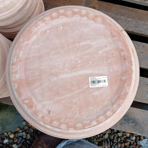 May include: A collection of terracotta-coloured, round plant saucers. Each saucer features a raised edge with a decorative pattern. The saucers are made of a porous material, designed to catch excess water from potted plants.