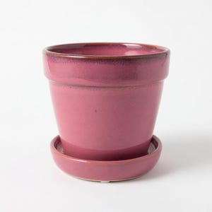 May include: A deep pink ceramic plant pot with a matching circular saucer. The pot features a smooth, glossy finish with a slightly darker, brownish-purple rim at the top edge. The design is simple and functional, with a tapered shape and a wide opening.