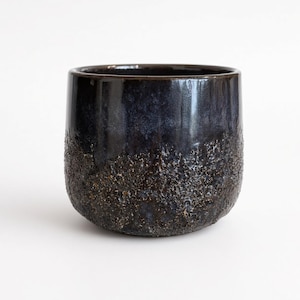 May include: A cylindrical ceramic cup featuring a dual-texture finish. The upper portion has a smooth, glossy dark blue and black glaze, while the lower section displays a rough, cratered, and textured surface in shades of dark brown and black.
