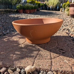 May include: A round, terracotta-coloured planter bowl with a wide opening and a raised base. The bowl is on a brick patio with small rocks in the foreground. Plants are visible in the background.