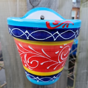 May include: A colourful ceramic wall-mounted plant pot. The pot is decorated with bands of blue, red, yellow, and white, with white scroll patterns. It's designed to be hung on a wall or fence, adding a bright touch to a garden or patio.