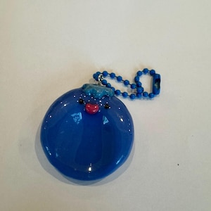 May include: A round, glossy blue charm featuring a small, textured blue top, two tiny black dot eyes, and a small pink heart-shaped mouth. The charm is attached to a matching blue ball chain, creating a circular pendant design with a smooth finish.