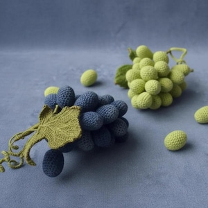 May include: Two clusters of crocheted grapes, one in dark blue and one in light green, each featuring a detailed green crocheted leaf and stem. Several individual crocheted grape spheres in matching colours are scattered around the main clusters.