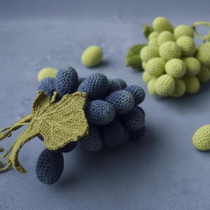 Könnte beinhalten: Gehäkelte Trauben in Blau- und Grüntönen, mit detailliertem Blatt und Stiel. Die handgefertigten Früchte sind vor einem weichen blauen Hintergrund platziert, der die Textur und Handwerkskunst des Garns hervorhebt.