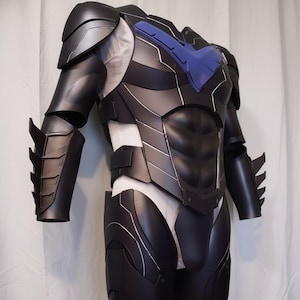 Può includere: Un costume da supereroe nero e grigio con dettagli blu. Il costume presenta spalline corazzate, protezioni per le braccia e un'armatura pettorale. Il design include finiture argentate e un'emblema blu sul petto.