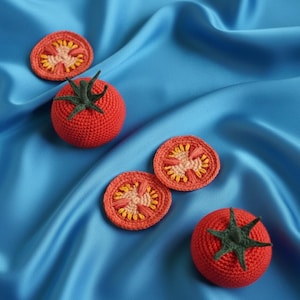 Könnte beinhalten: Gehäkelte Tomaten und Tomatenscheiben in Rot, Orange und Grün. Die Tomaten sind auf einem blauen Satinhintergrund platziert. Die Tomaten haben grüne Stiele, und die Scheiben zeigen die inneren Details.