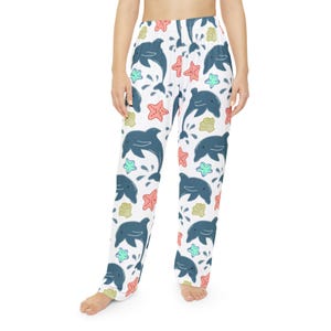 May include: White pajama pants with a repeating pattern of blue dolphins, red starfish, and green and yellow seashells. The pants have a relaxed fit and are made of a soft material.