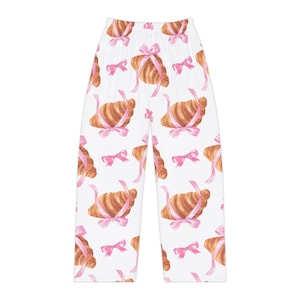 Könnte beinhalten: Weiße Schlafanzughose mit einem sich wiederholenden Muster aus Aquarell-Croissants, die mit rosa Bändern und Schleifen gebunden sind. Die Hose hat eine lockere Passform und ein bequemes Design.