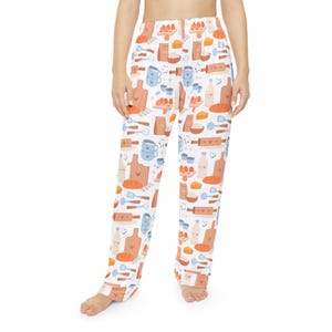 May include: White pajama pants with a repeating pattern of baking-themed illustrations. The design includes images of rolling pins, cutting boards, eggs, bread, and milk bottles in shades of orange, blue, and brown.