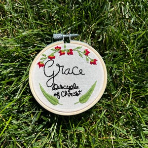 May include: Embroidered hoop art with a white background, featuring the name "Grace" in cursive script and the phrase "Disciple of Christ" in smaller script. The hoop is decorated with green leaves and red flowers.