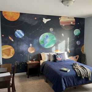 May include: A bedroom with a space-themed mural featuring planets, rockets, and astronauts. The room includes a bed with a navy blue comforter, pillows, and a desk with a laptop and model rocket. A skateboard leans against the wall.