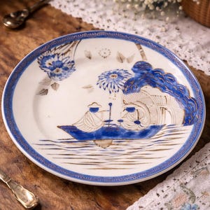 May include: A white ceramic plate with a blue and gold design. The plate features a blue boat scene with figures and floral accents. The rim has a blue and gold border. Decorative tableware.