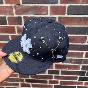 Könnte beinhalten: Schwarze Baseballkappe mit einem weißen gestickten Logo und Perlenverzierungen. Die Kappe hat eine Kette mit einem Kreuzanhänger.