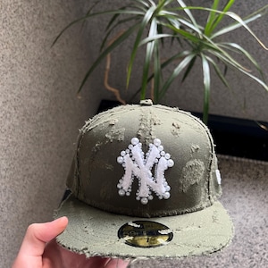 Könnte beinhalten: Eine grüne, abgenutzte Baseballkappe mit einem weißen, gestickten New York Yankees-Logo, das mit weißen Perlen verziert ist. Die Kappe hat einen gebogenen Schirm und ein goldenes New Era-Logo.