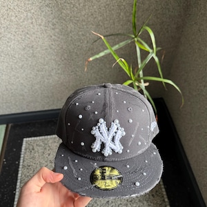 Könnte beinhalten: Graue Baseballkappe mit weiß gesticktem New York Yankees Logo. Die Kappe ist mit kleinen weißen Perlen verziert und hat einen ausgefransten Schirm. Der New Era Aufkleber ist sichtbar.
