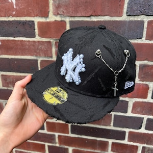 Könnte beinhalten: Schwarze Baseballkappe mit einem weißen gestickten New York Yankees Logo, das mit Perlen verziert ist. Die Kappe hat einen Used-Look und verfügt über eine silberne Kette mit einem Kreuzanhänger.