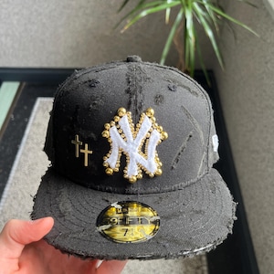 Könnte beinhalten: Schwarze, abgenutzte Baseballkappe mit einem weißen New York Yankees-Logo, das mit goldenen Perlen verziert ist, und einem goldenen Kreuz an der Seite. Die Kappe hat einen schwarzen verstellbaren Riemen mit einem goldenen '59FIFTY'-Etikett.