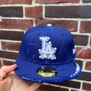 Può includere: Un cappellino da baseball blu con le lettere "LA" in perle bianche. La tesa e i bordi del cappellino hanno un aspetto invecchiato. Il cappellino è taglia 7 3/8.