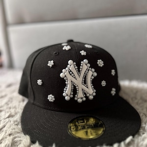 Könnte beinhalten: Eine schwarze Baseballkappe mit einem weißen New York Yankees Logo, das mit weißen Perlen verziert ist. Die Kappe hat einen schwarzen Schirm und ein goldenes New Era 59FIFTY Label.